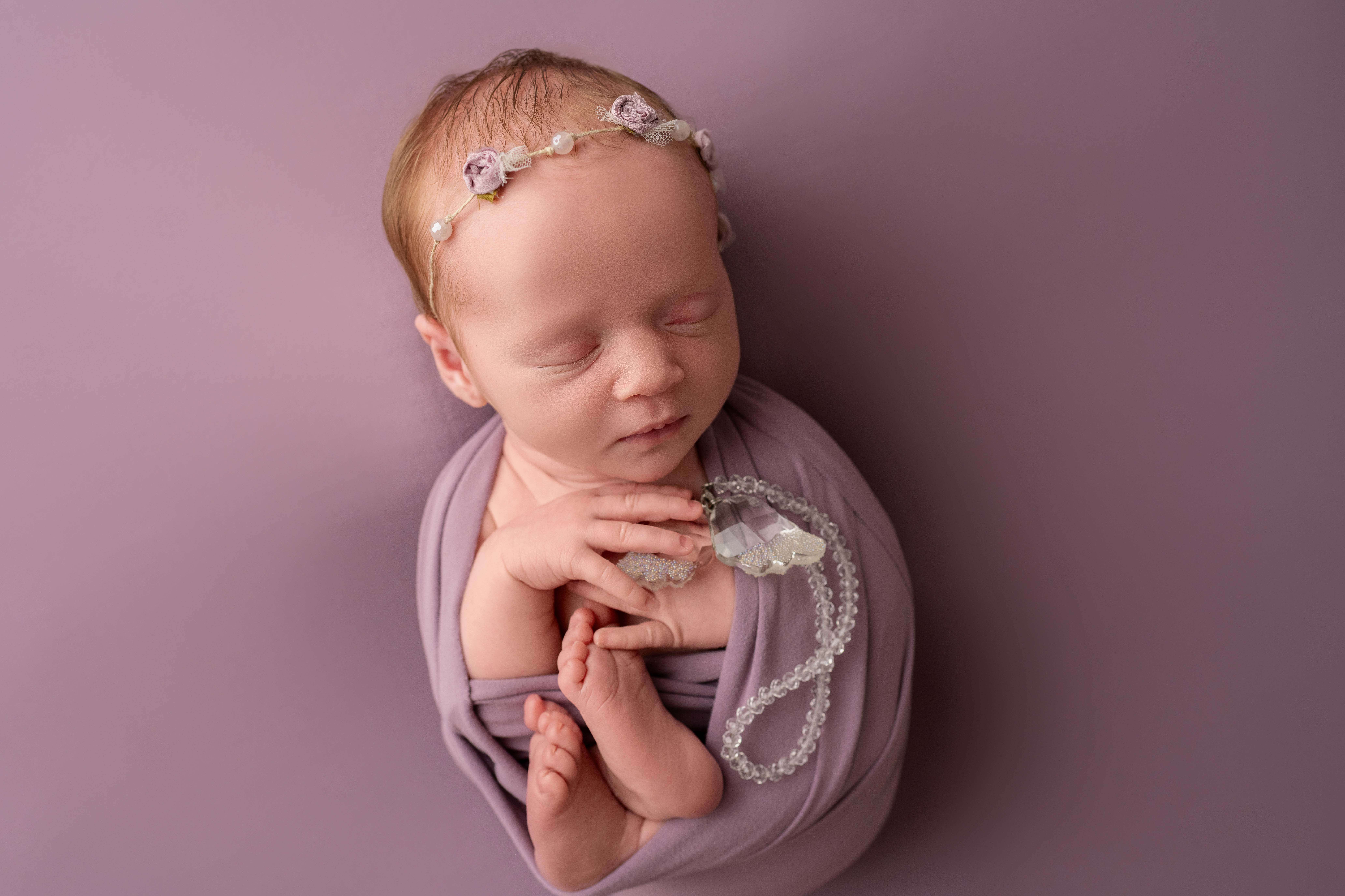 A sleeping newborn wrapped in a purple swaddle holding a bracelet after visiting baby boutiques in Kennewick, WA