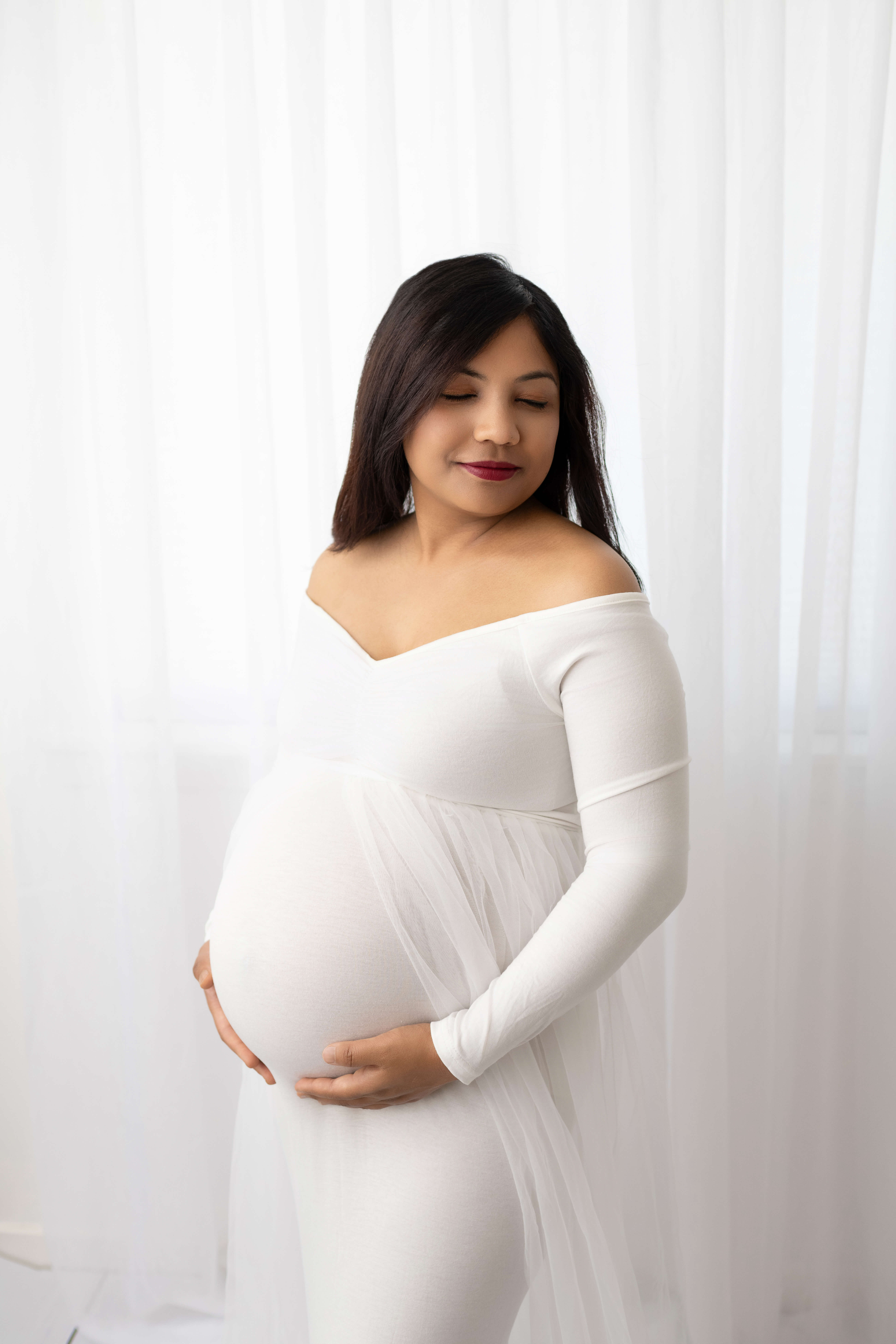A smiling pregnant woman in a white maternity gown smiles down her shoulder while standing in a studio after finding perfect doulas in Tri Cities, WA