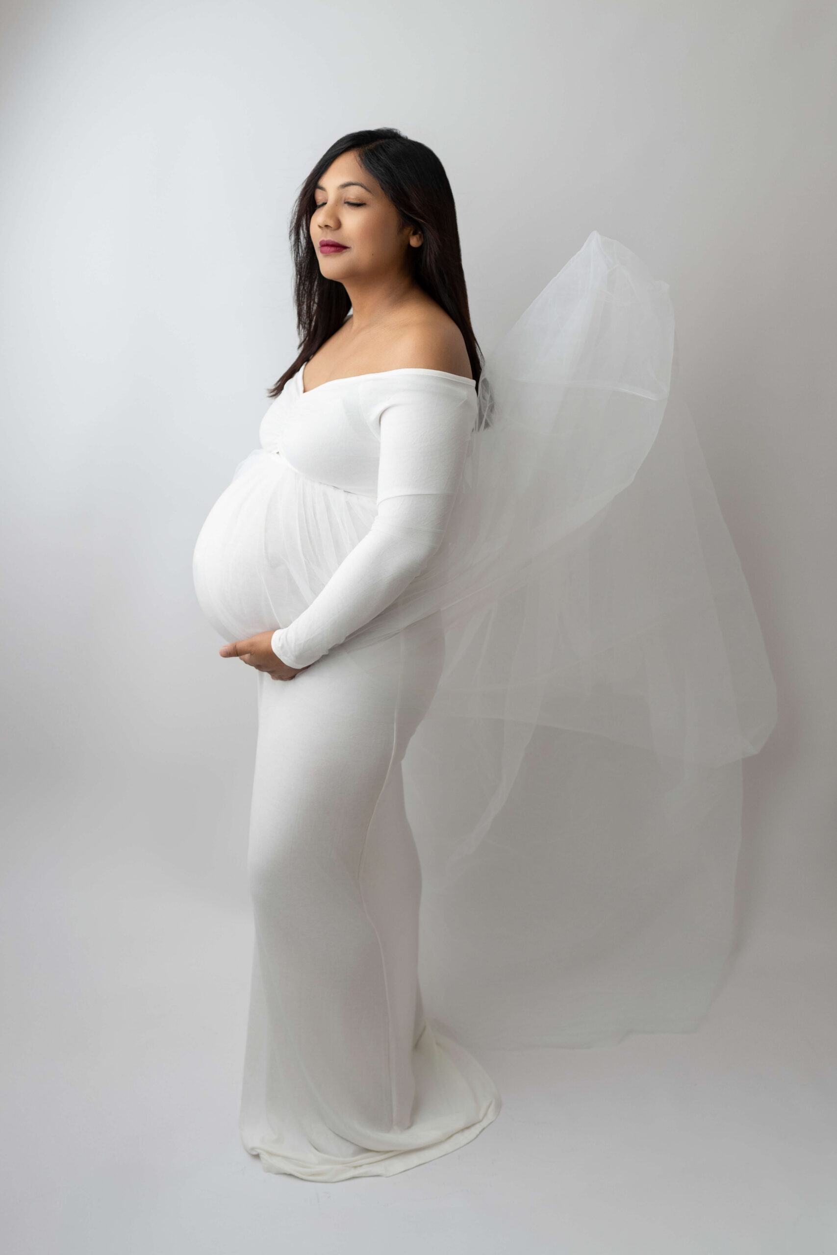 A mother to be stands in a white maternity gown in a studio with hands under her bump after meeting with doulas in Tri Cities, WA