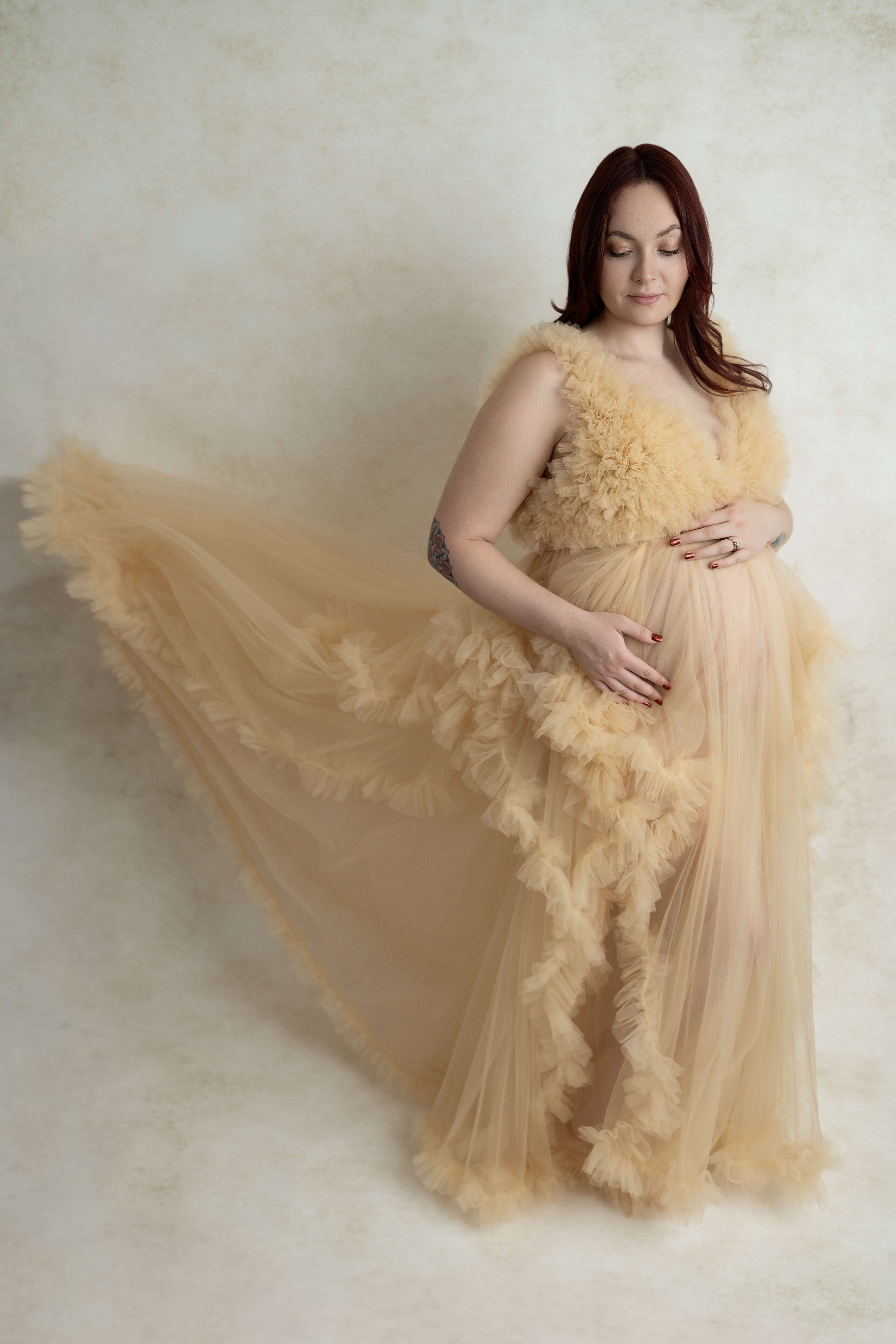 A pregnant woman smiles down to her bump as her yellow tule maternity gown train flows around her