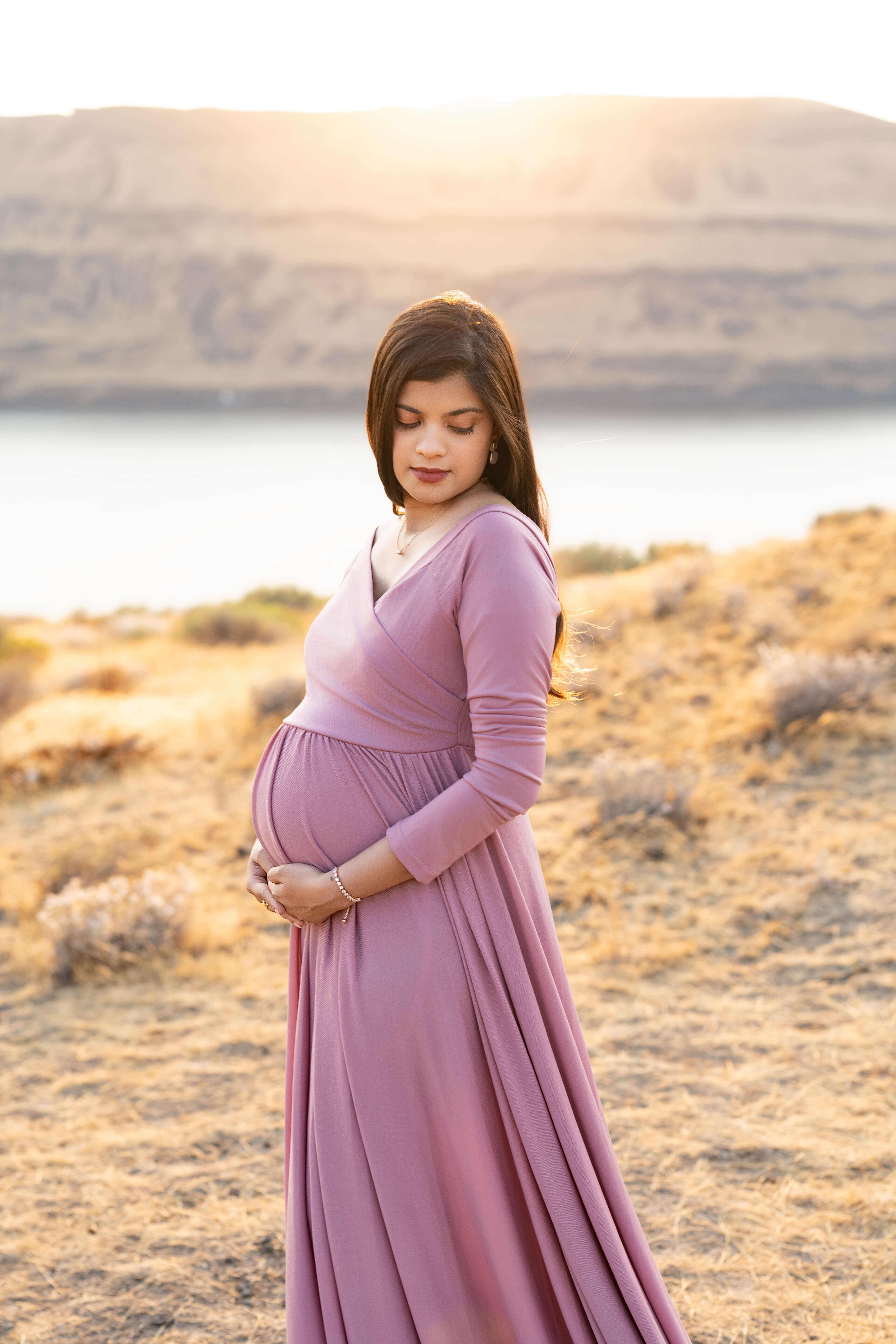 A mother to be stands on a cliff overlooking the ocean at sunset in a pink maternity gown while gazing down her shoulder after enjoying a 3d ultrasound in tri cities, WA