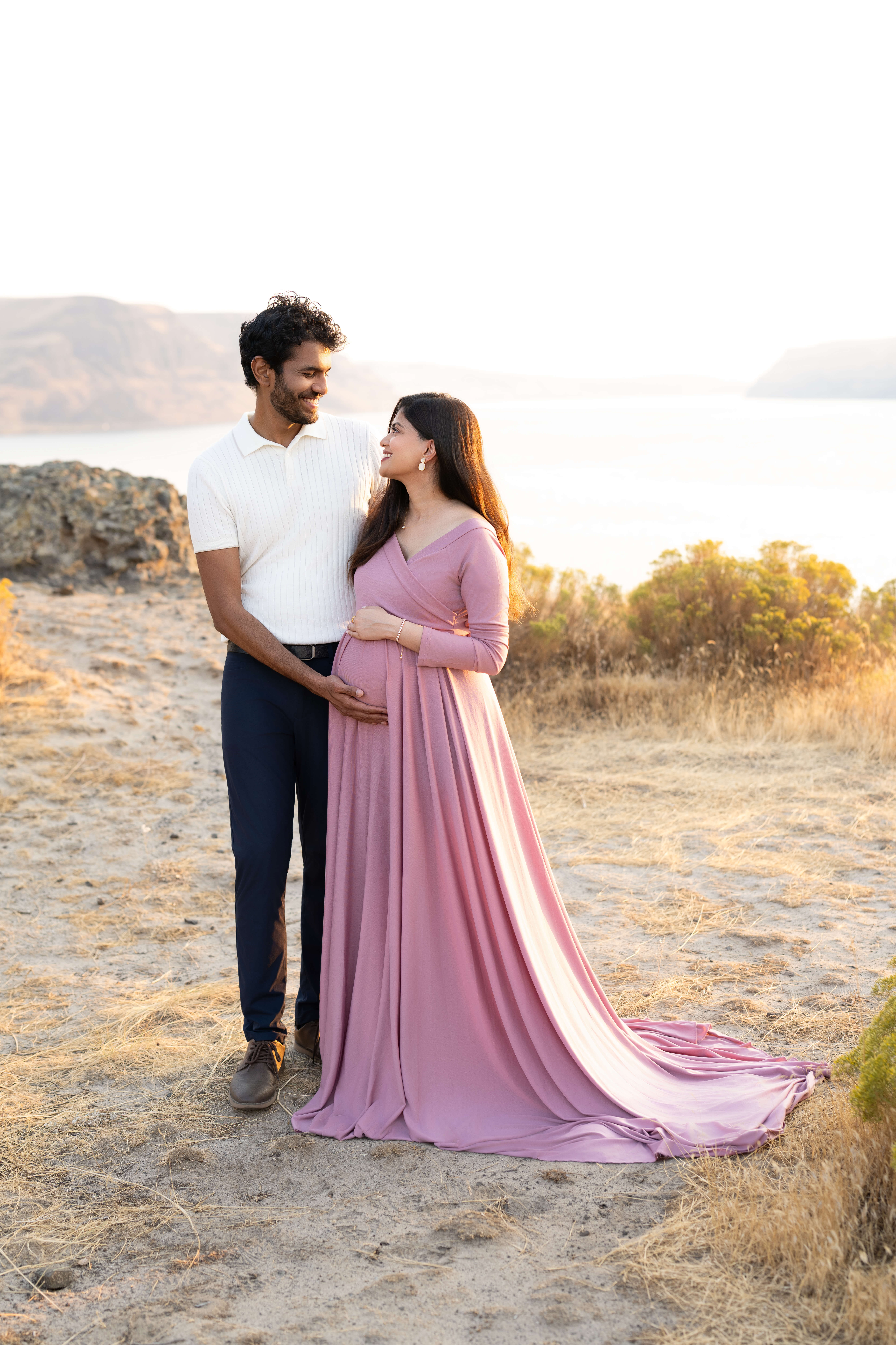 A happy expecting couple stands on a cliffside at sunset in pink and white smiling at each other overlooking the water after finding a 3d ultrasound in tri cities, WA