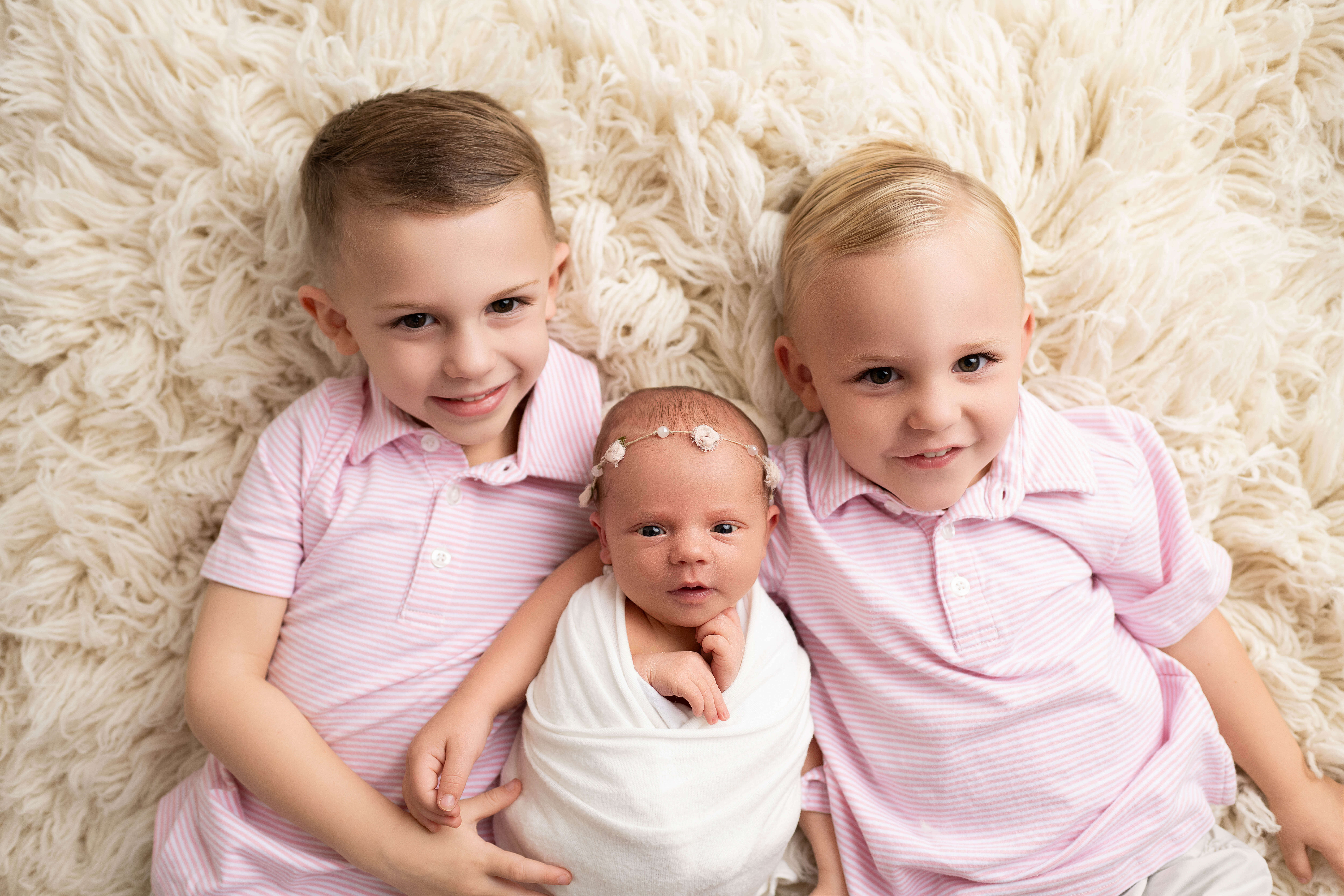 Toddler brothers in matching pink polos lay hugging their newborn baby sister in a white swaddle