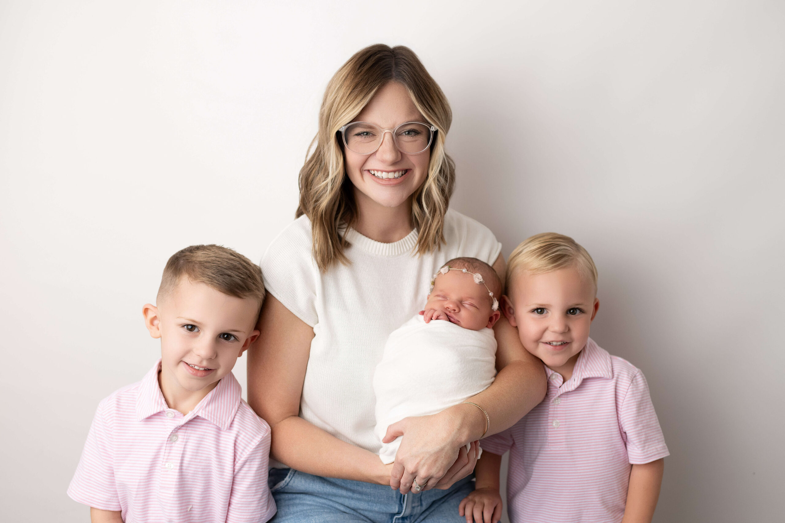 A happy mom sits with her toddler sons in matching pink polos while holding a newborn in a studio after meeting a nanny in tri-cities, WA