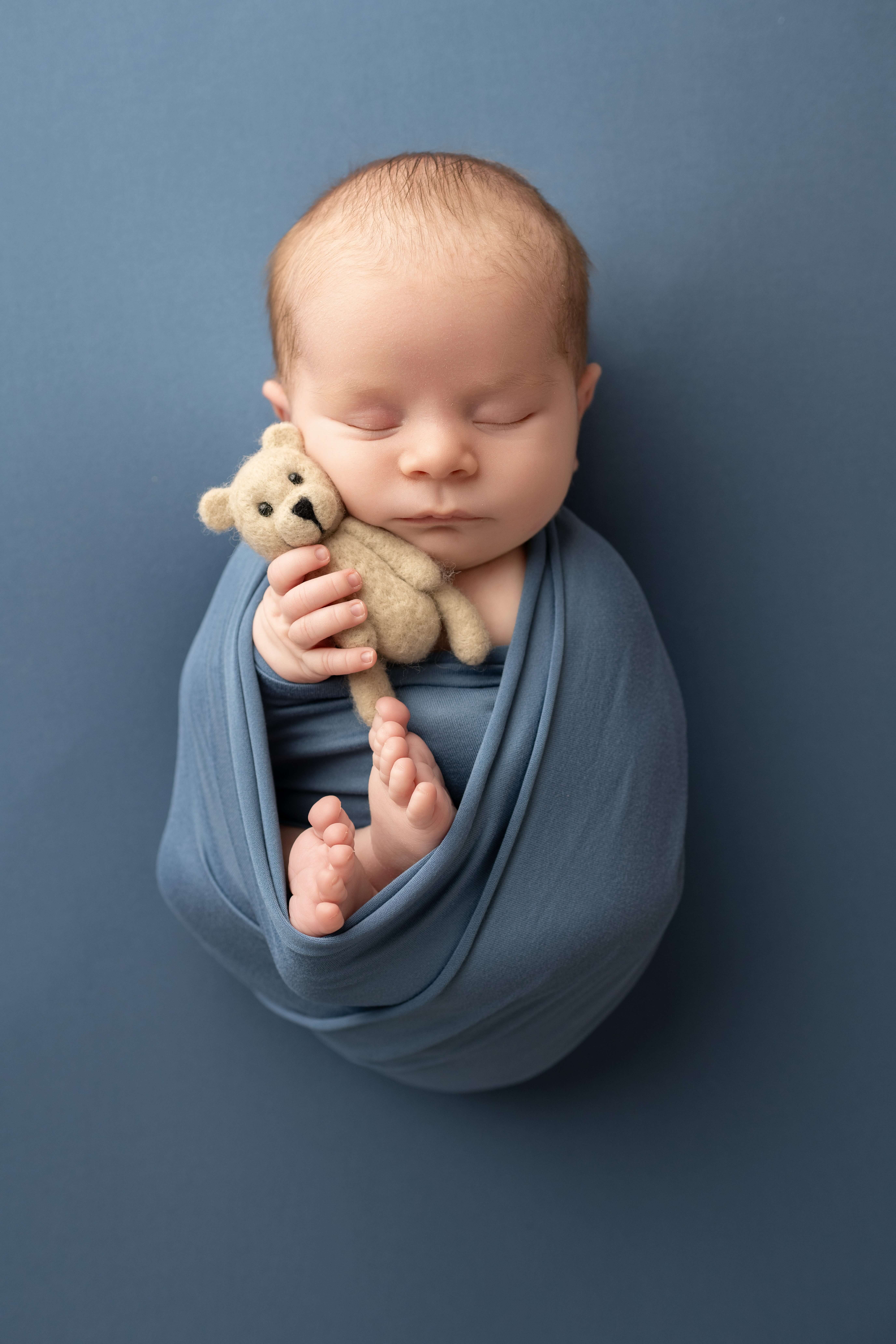 A newborn baby sleeps in a blue swaddle holding a tiny bear against his cheek thanks to lactation consultants in tri-cities